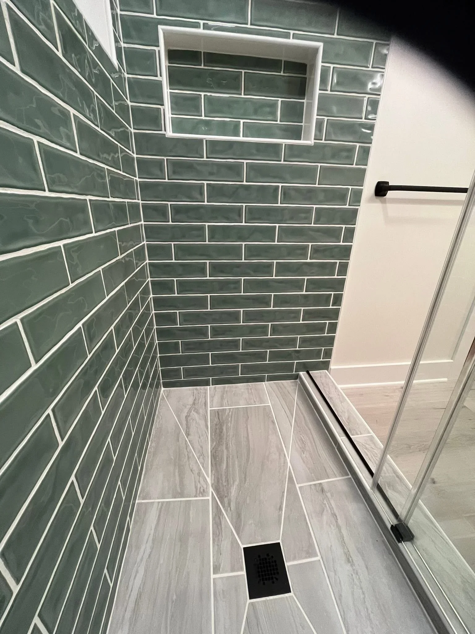 Sage green subway tile shower with herringbone wood-look floor and frameless glass door